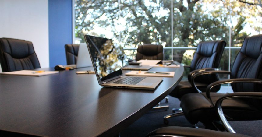 Sala de reuniões moderna com mesa de madeira, laptop e cadeiras confortáveis, iluminação natural abundante.