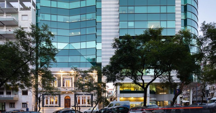 Cena urbana com edifício moderno em vidro e prédio tradicional ornado, carros e pedestres.